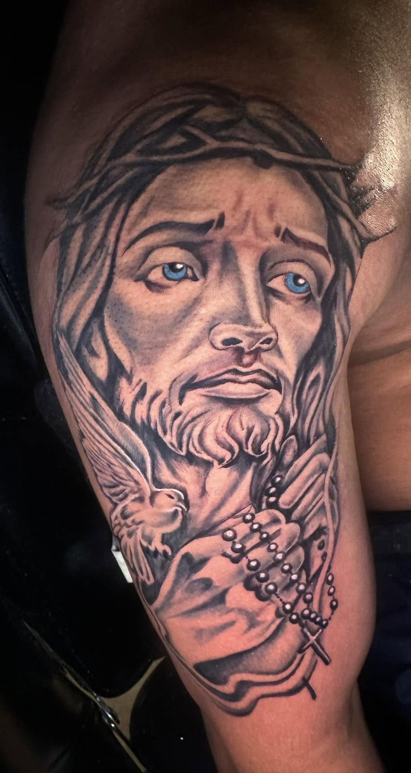Black and grey tattoo of Jesus wearing a crown of thorns with blue eyes, hands clasped around a rosary, on an upper arm.